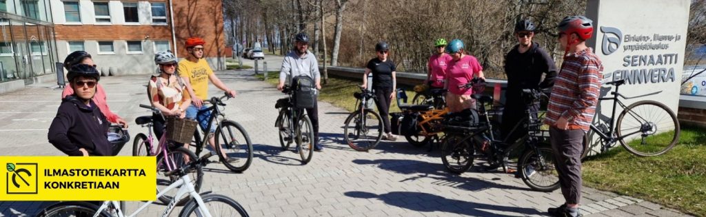 Ihmisiä seisoo polkupyöriensä kanssa toimiston pihalla kevätpäivänä.