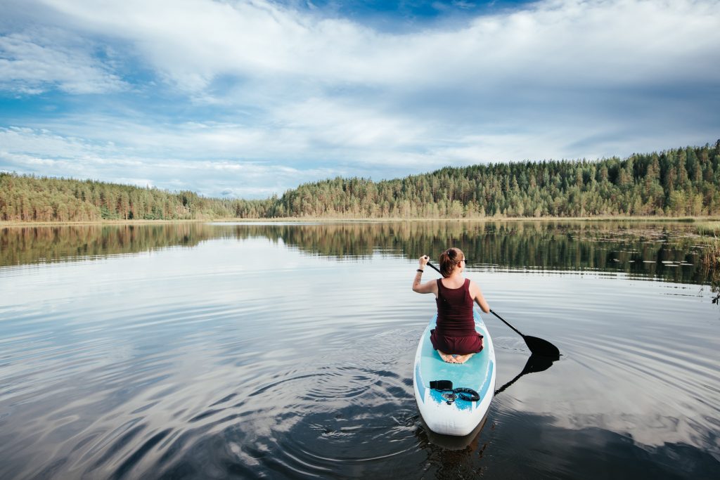 Henkilö on polvillaan SUP-laudan päällä ja meloo kevyen näköisesti. Suppailijan ympärillä on kaunis järvi- ja metsämaisema kesäaikaan kuvattuna.