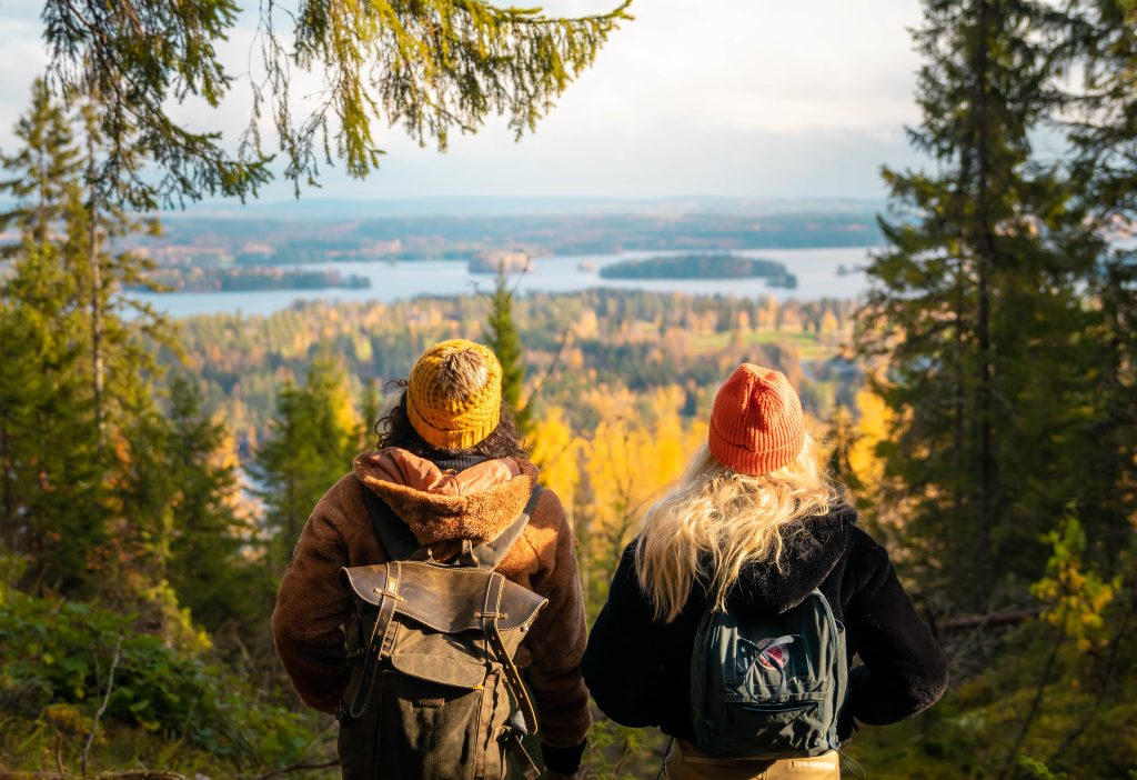 Kaksi henkilöä seisoo selkä kameraa kohti syksyisessä metsä- ja järvimaisemassa.