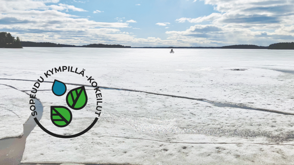 Kuvassa Sopeudu kympillä kokeiluiden tunnus ja taustakuvana pohjoissavolainen järvimaisema kevään jäiden lähdön aikaan ja pyöräilijä jäällä mustana siluettina horisontissa