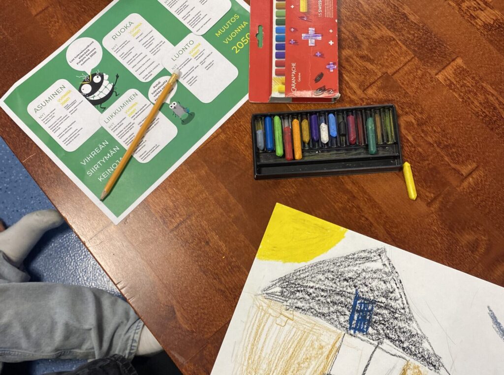 Kuvituskuvassa puinen ruskea pöytä, jonka päällä väriliituja ja piirustus.