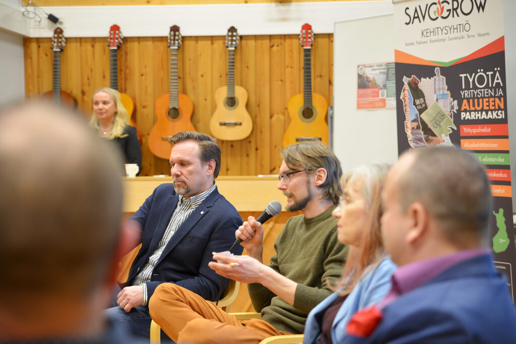 Henkilöt istuvat penkeillä, yksi puhuu mikrofoniin.