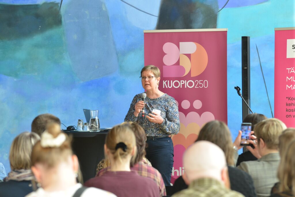 Kansanedustaja Krista Mikkonen seisoo yleisön edessä ja puhuu mikrofoniin.