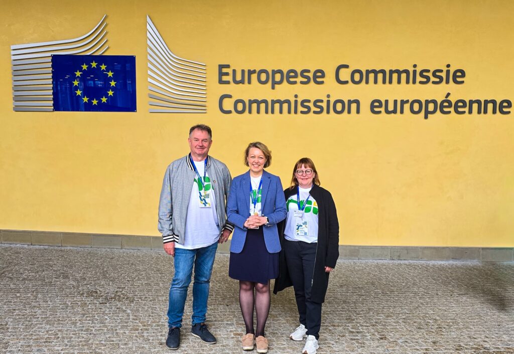 Kolme henkilöä seisovat EU komission päärakennuksen edessä ja hymyilevät.